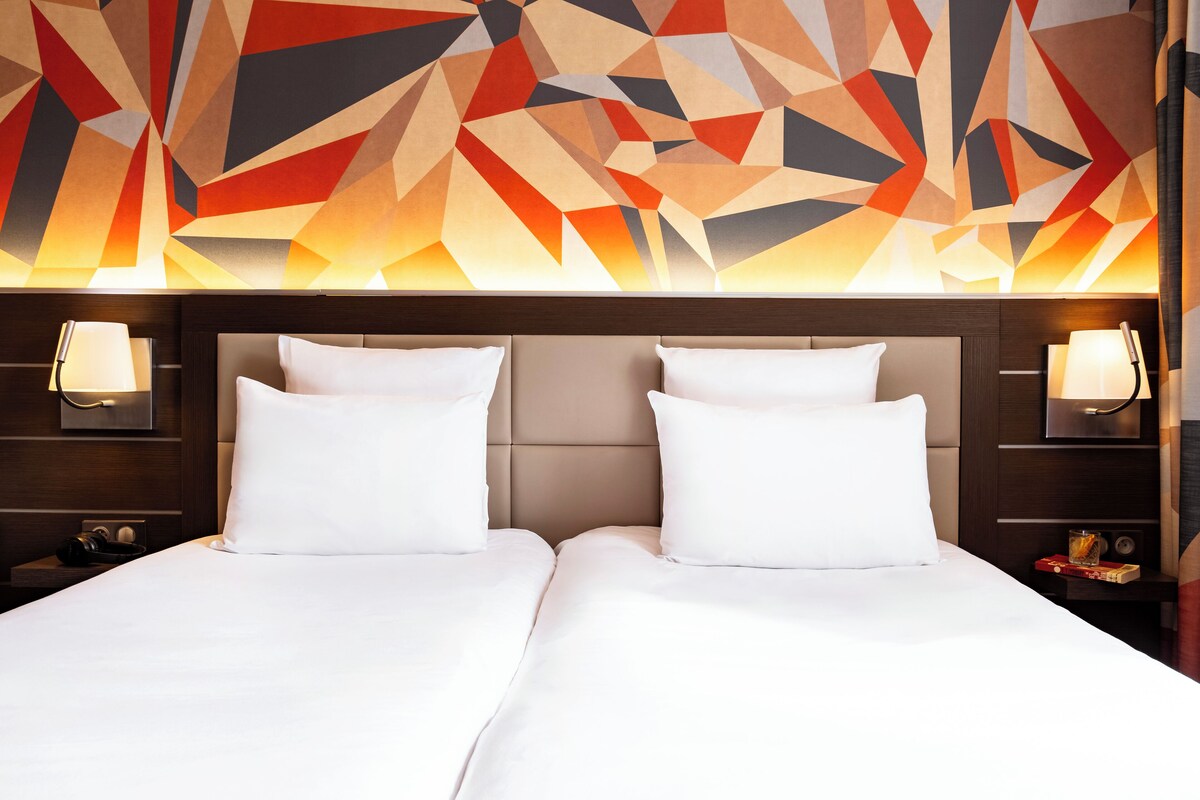 A hotel room with a modern geometric wall design, two beds with white linens, and bedside lamps.