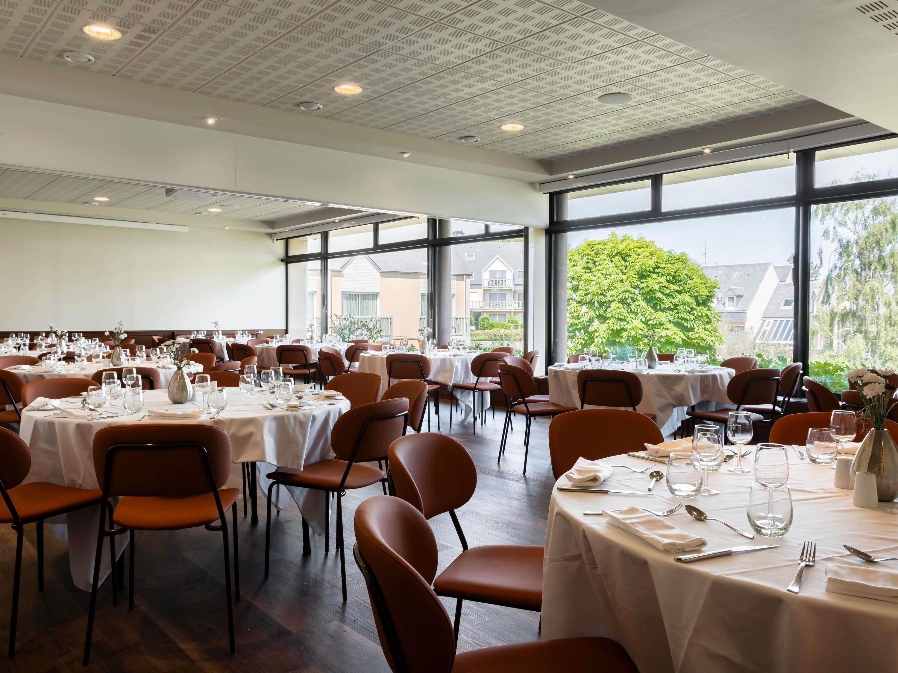 A large dining hall with multiple tables set for dining, large windows, and a high ceiling with ventilation.