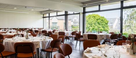 A large dining hall with multiple tables set for dining, large windows, and a high ceiling with ventilation.