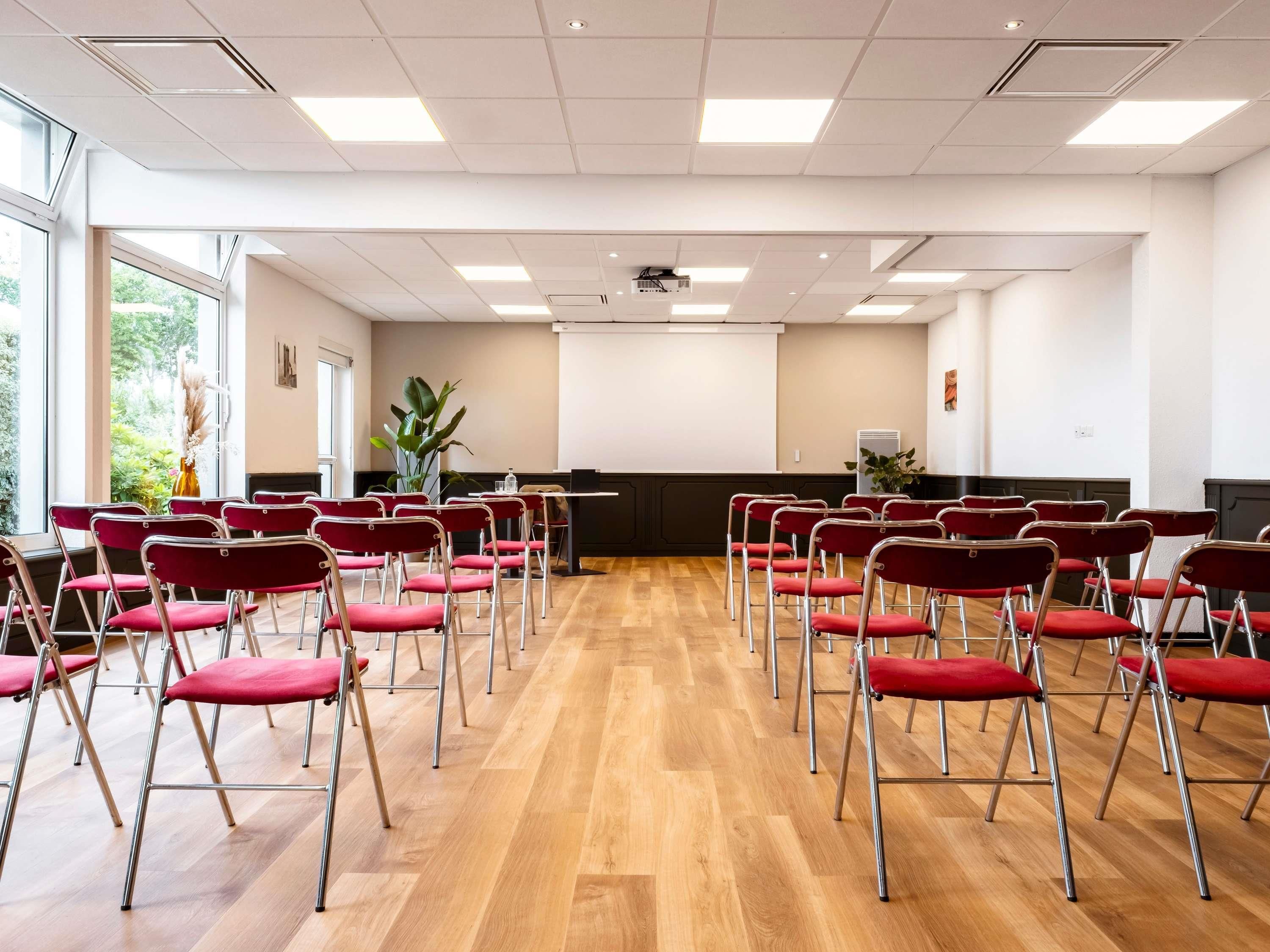 Une salle de conférence avec des rangées de chaises rouges, un projecteur et un tableau blanc.
