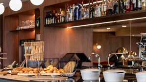 Zona de bar con estantes de botellas de licores, una máquina de café y un bufé con pan y bollería.
