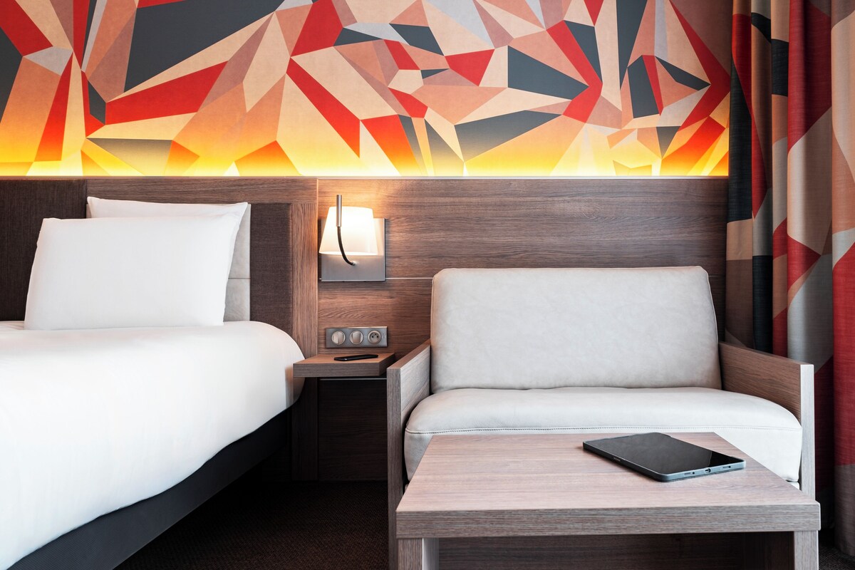 A hotel room with a bed, a sofa, a nightstand with a lamp, and a tablet on a table.