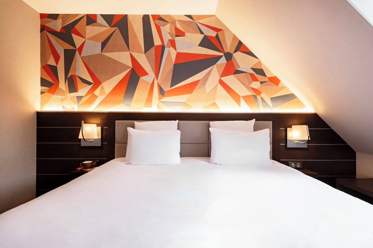 A modern hotel room with a large bed, two bedside lamps, and a geometric wall art above the bed.