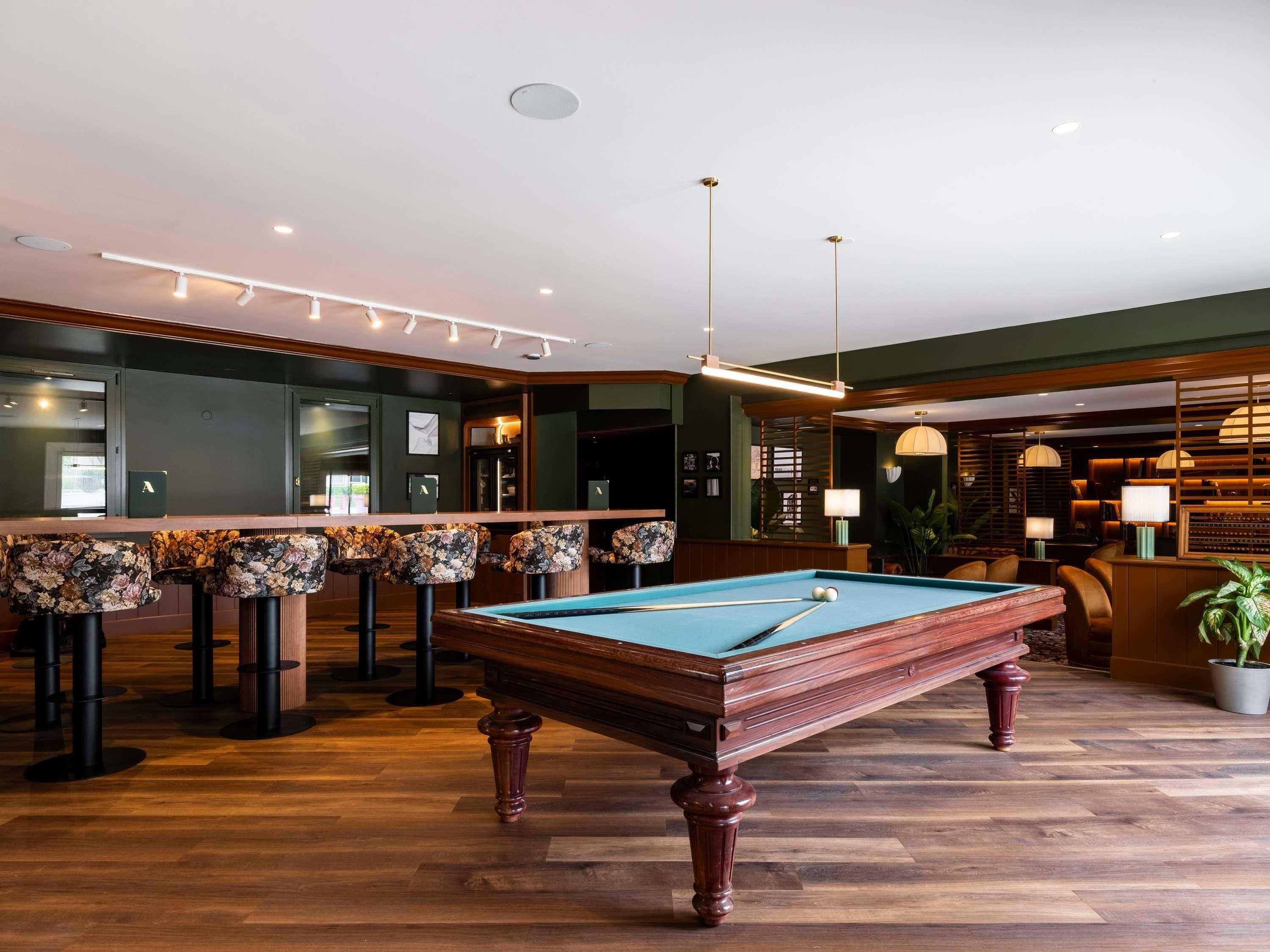 A pool table in a room with wooden flooring, bar stools with floral upholstery, and a bar area with a green wall and wooden accents.