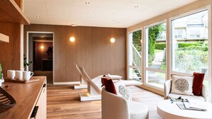 Una habitación de hotel moderna con suelo de madera, un sofá blanco, una mesa redonda y ventanas amplias con vistas al exterior.