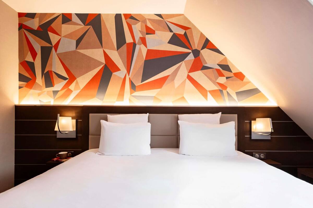 A hotel room with a modern geometric wall design, a large bed with white linens, and two bedside lamps.