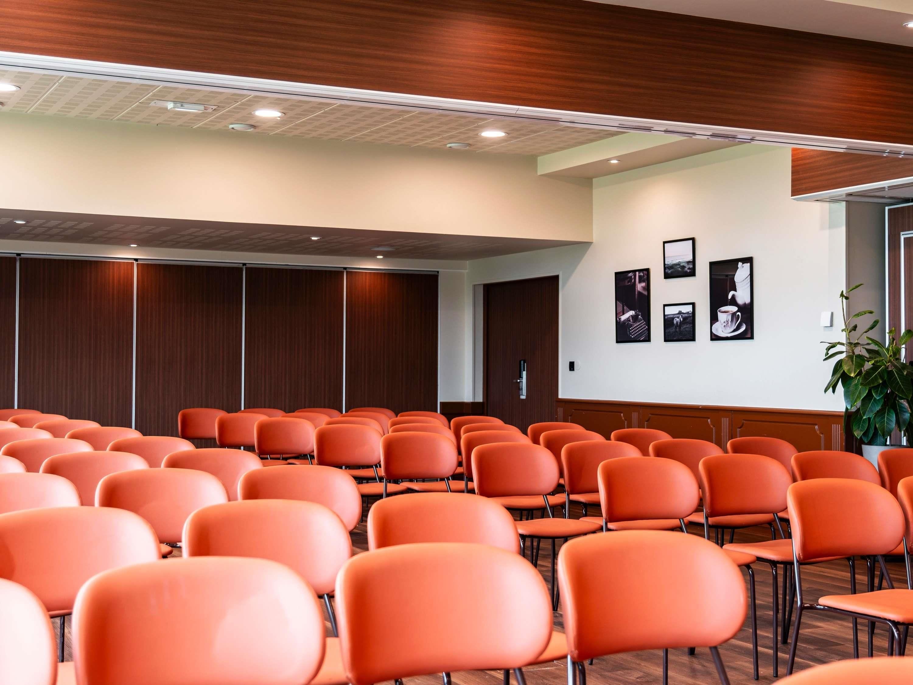 A conference room with rows of orange chairs, wooden paneling on the walls, and a door at the back.