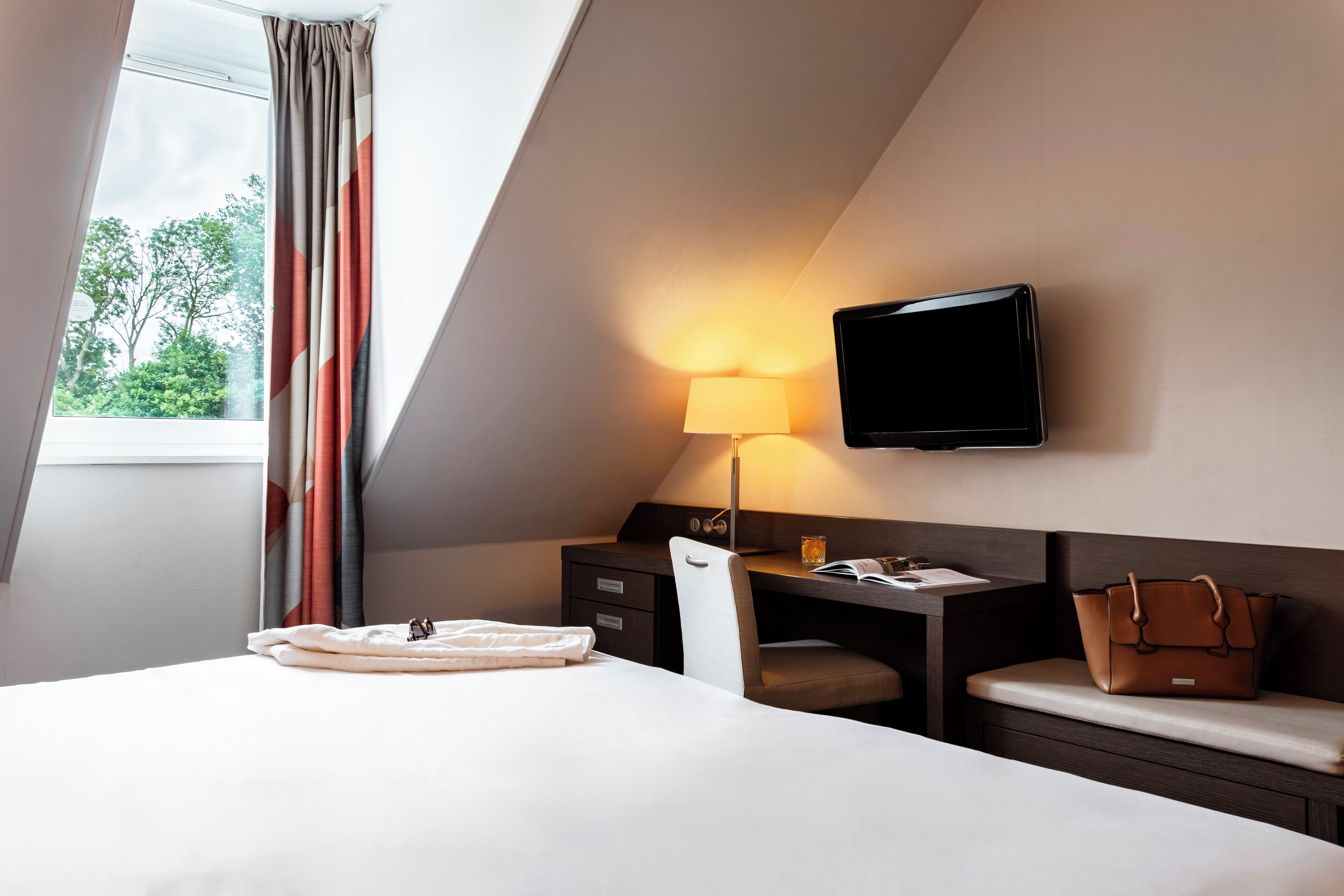 A hotel room with a bed, a desk with a lamp, a TV, and a window with curtains.