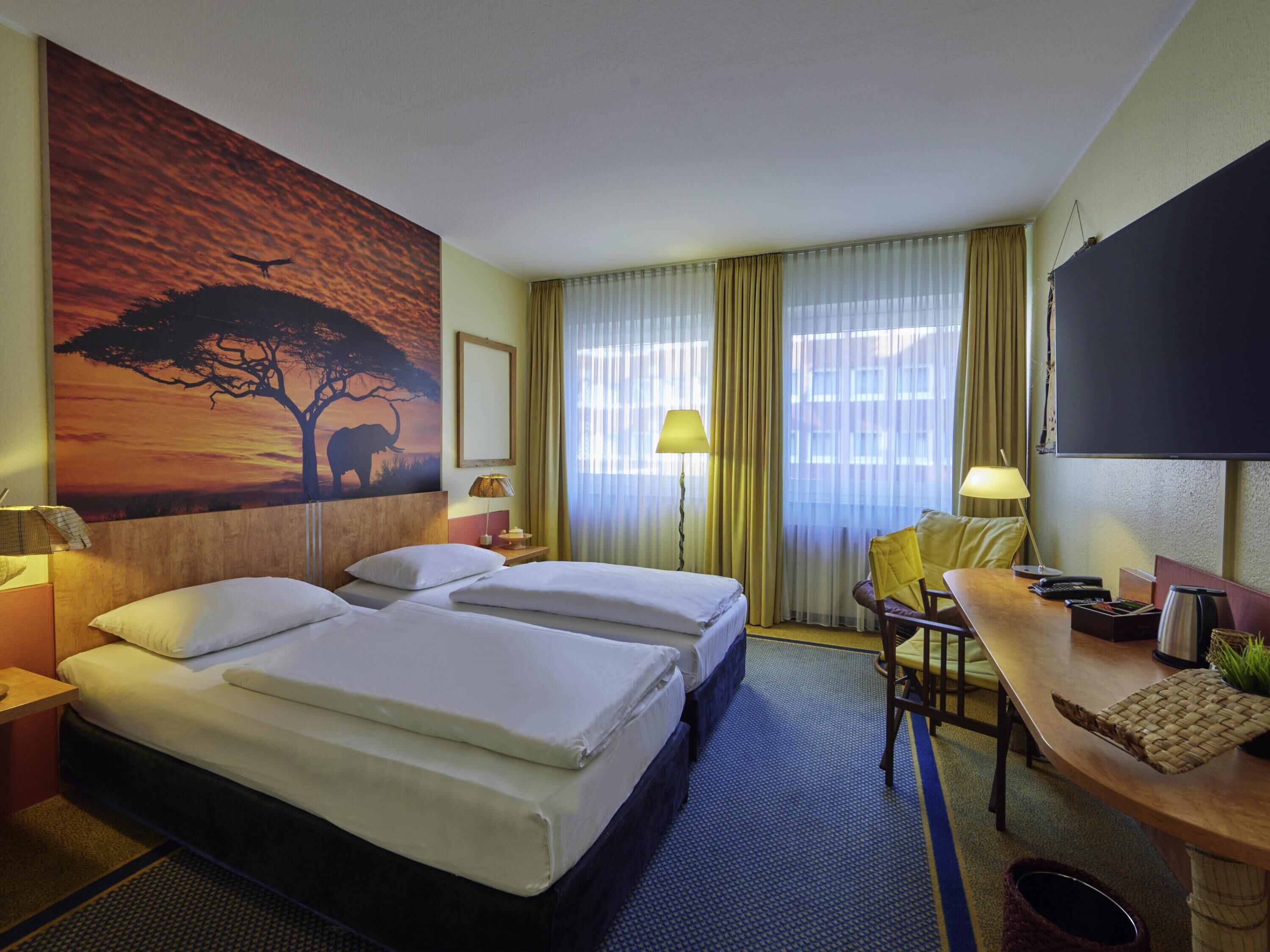 A hotel room with two beds, a desk, a chair, a TV, and a painting of a tree and elephant.