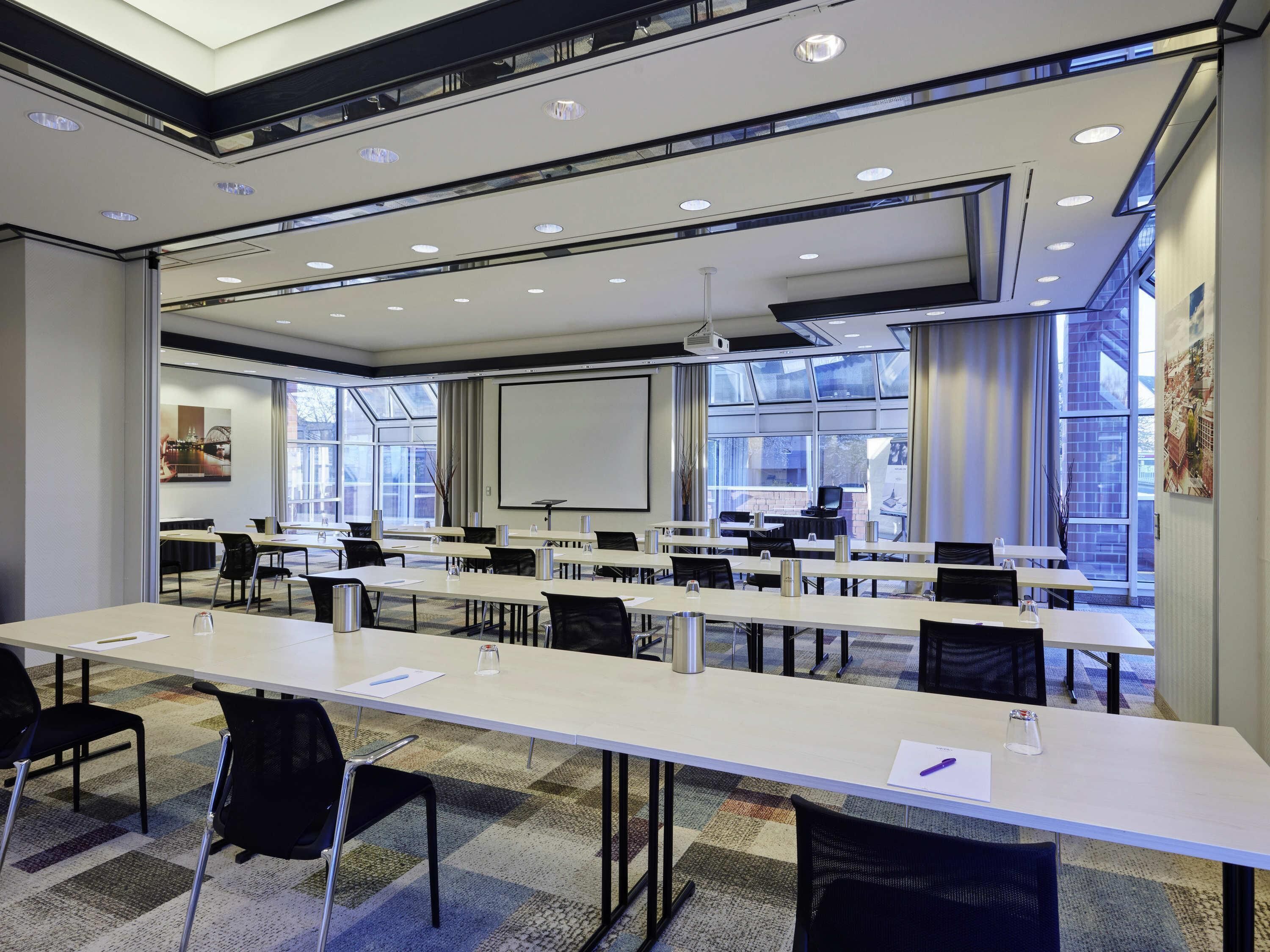 A conference room with long tables, chairs, a projector screen, and large windows.