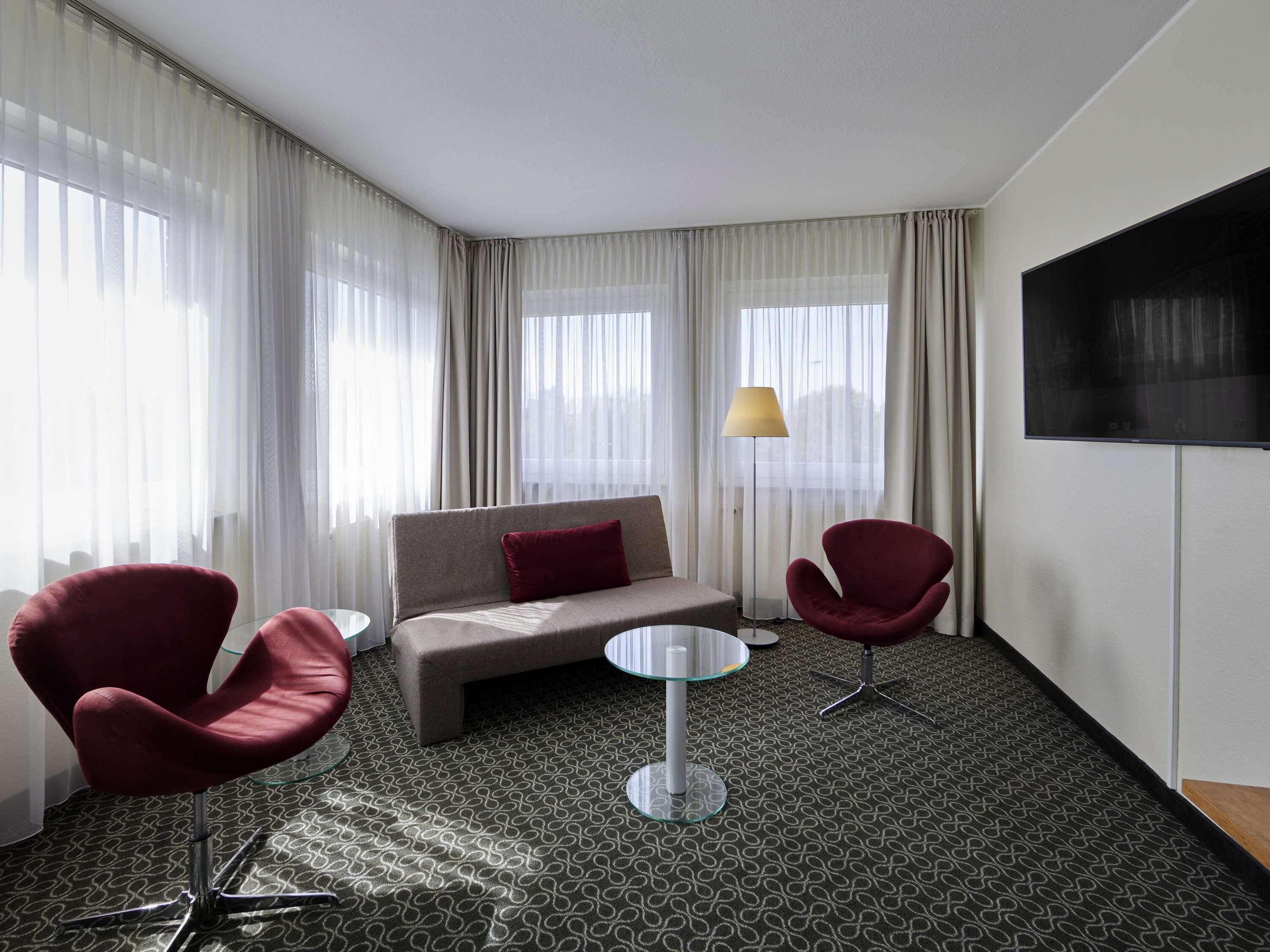 A hotel room with a sofa, two armchairs, a small table, a TV, and large windows with sheer curtains.