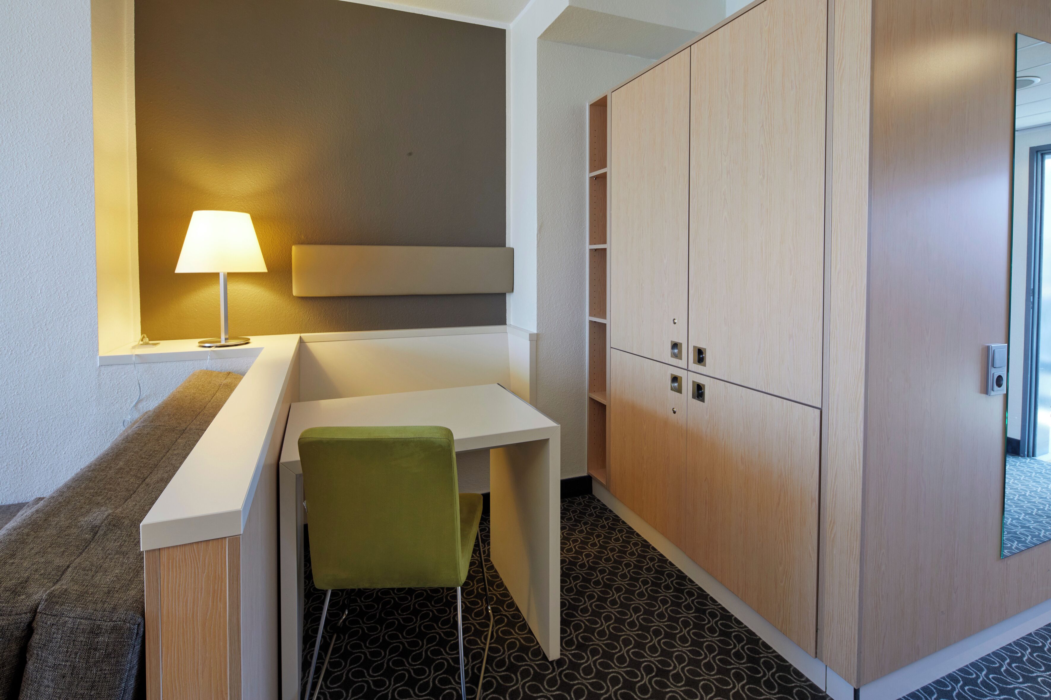 A small, modern room with a desk, a green chair, a lamp, and built-in wooden cabinets.