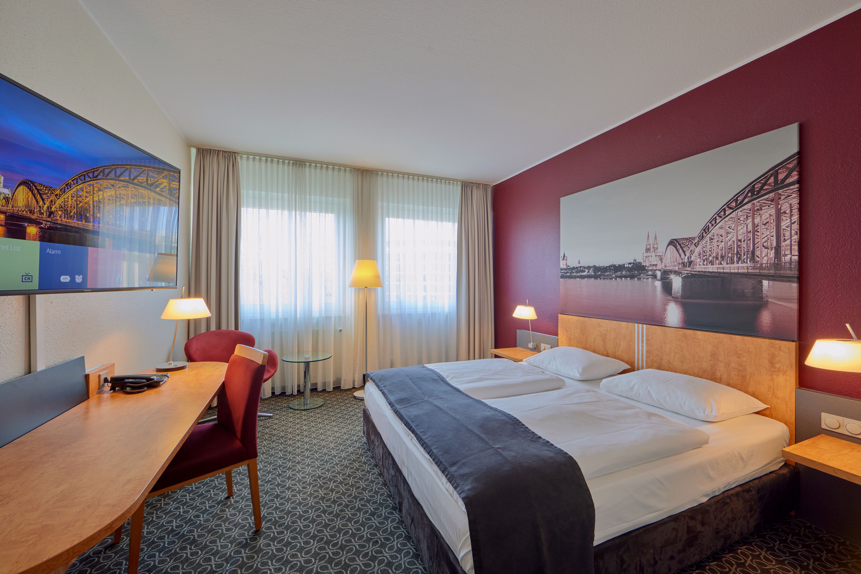 Quarto de hotel com uma cama grande, uma escrivaninha, uma cadeira, uma televisão e um quadro de uma ponte.