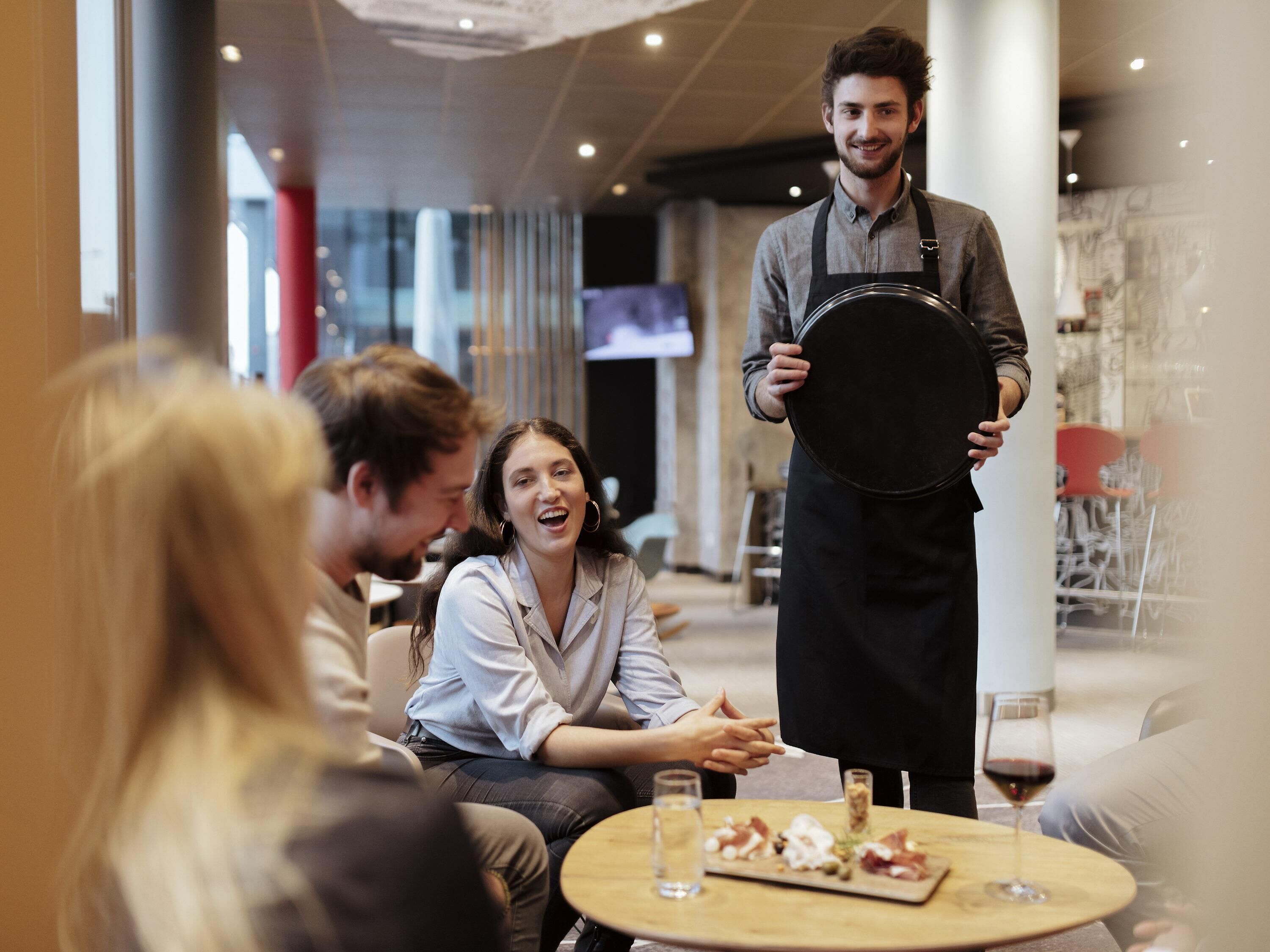 A restaurant interior with a round table, a waiter holding a tray, and a glass of red wine on the table.