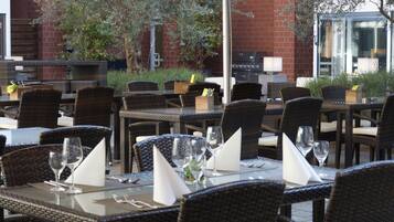 An outdoor dining area with wicker furniture and glass tables, featuring a brick wall and greenery in the background.
