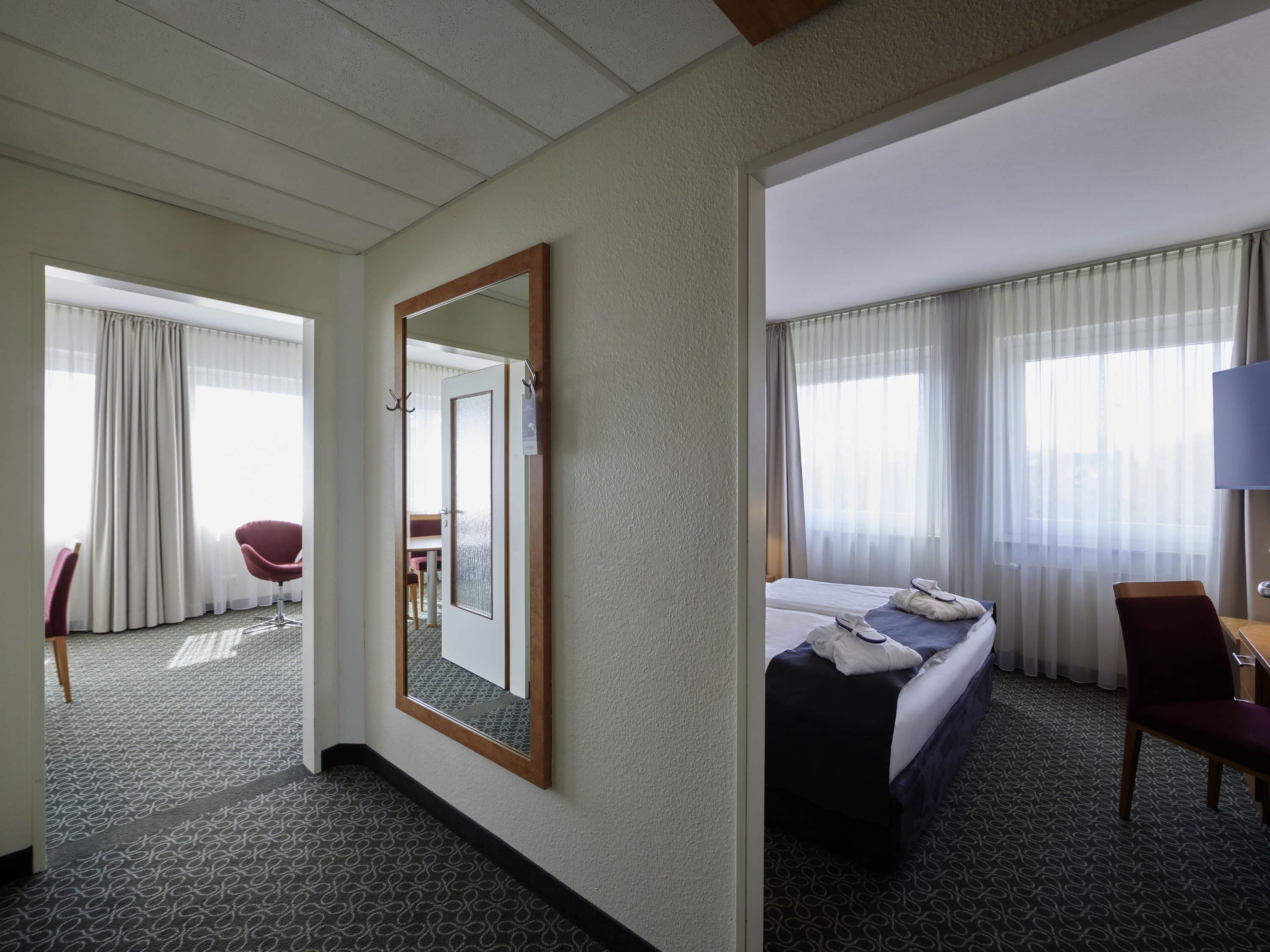 A hotel room with a bed, a desk, a chair, a mirror, and a view of the outside through large windows.