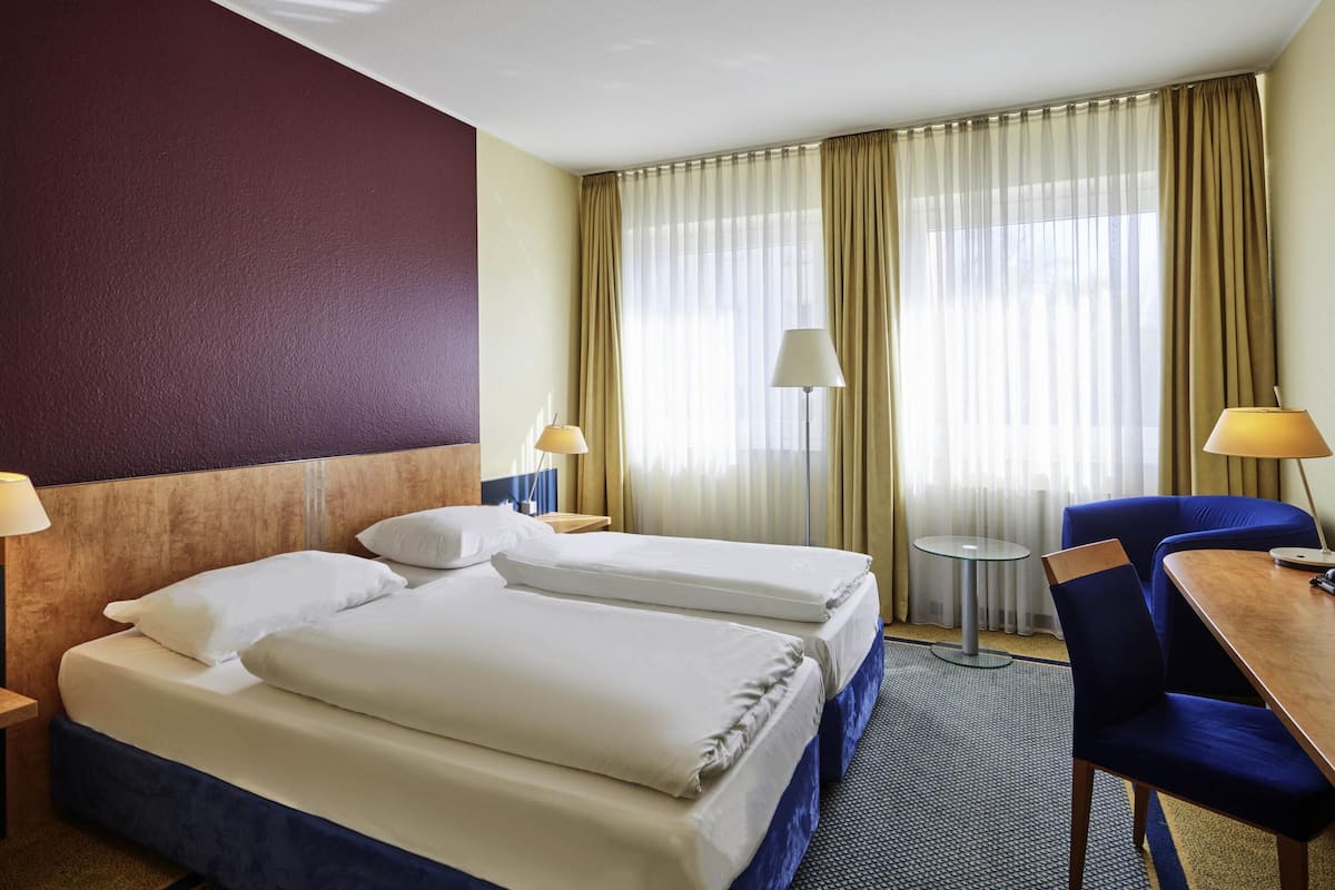 Een hotelkamer met twee bedden, een bureau, een stoel, een lamp en uitzicht op het omliggende landschap door dunne gordijnen.