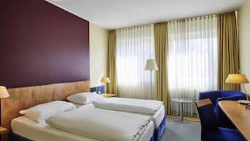 Camera d'albergo con due letti, una scrivania, una sedia, una lampada e vista all'esterno attraverso tende leggere.