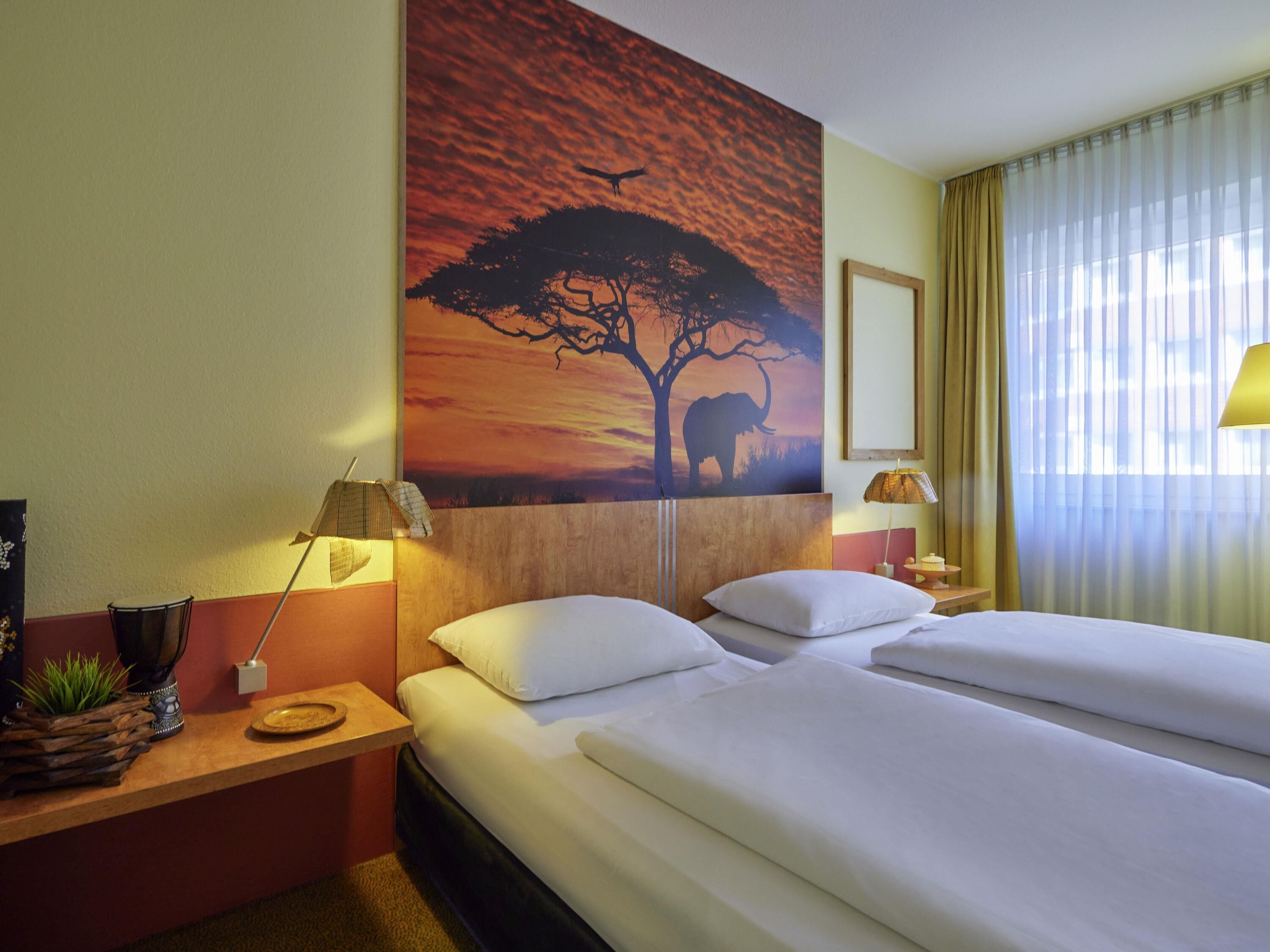 A hotel room with two beds, a painting of an elephant and tree, a lamp, and a nightstand with a plant.