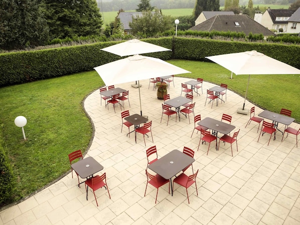 An outdoor patio with tables and chairs, surrounded by a hedge and houses in the background.