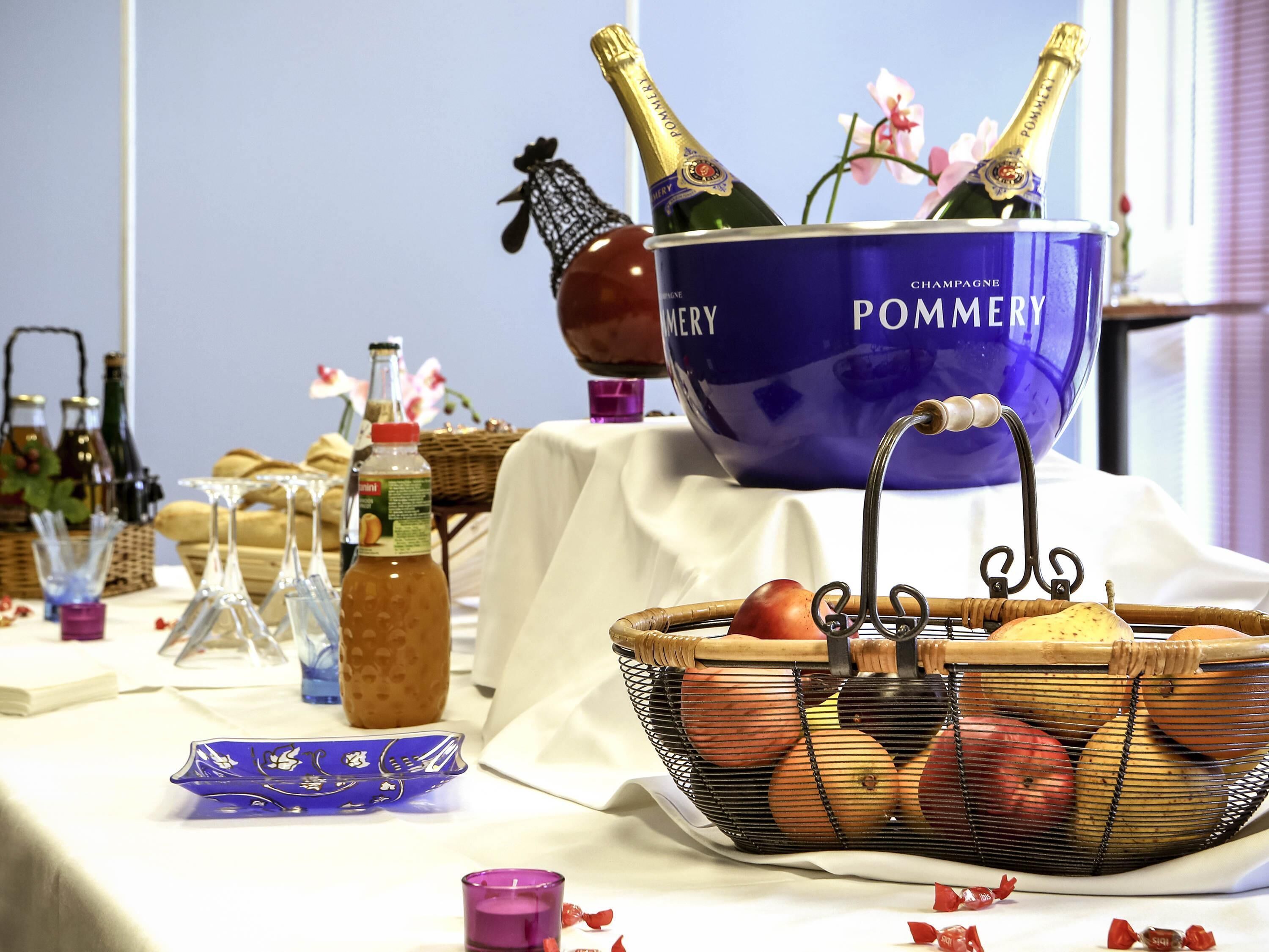 Una mesa con un tazón azul que dice 'POMMERY CHAMPAGNE', botellas, una cesta de frutas y diferentes botellas y copas.
