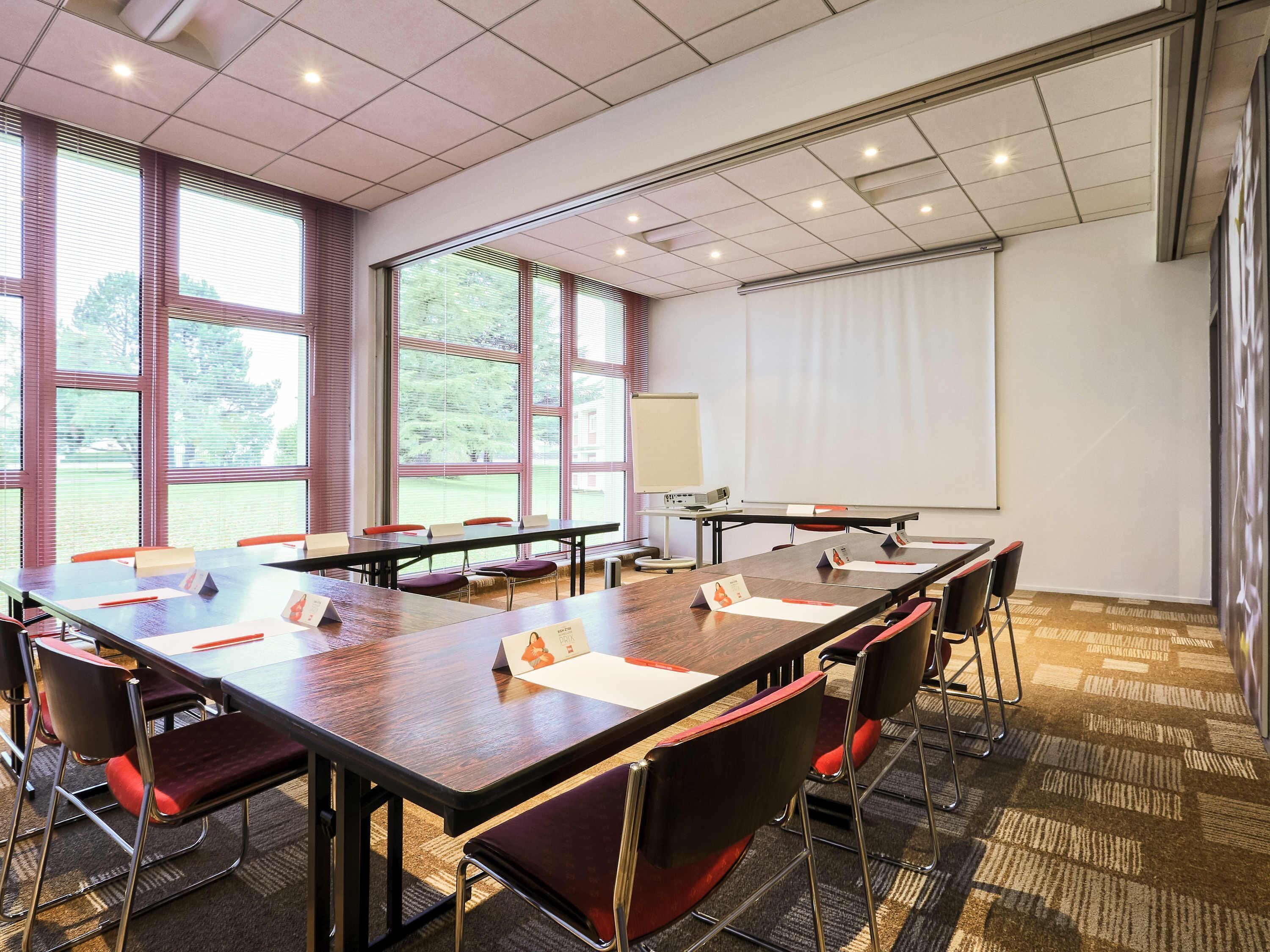 Une salle de conférence équipée d’une longue table, de chaises, d’un écran de projection et de grandes fenêtres.