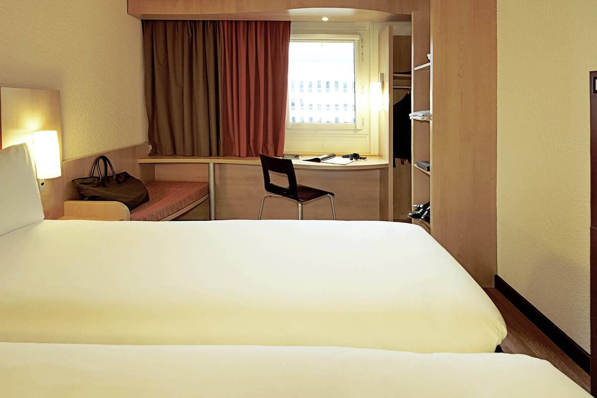 A hotel room with a large bed, a desk with a chair, a window with curtains, and a wardrobe.