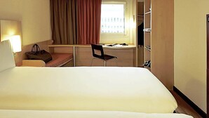 A hotel room with a large bed, a desk with a chair, a window with curtains, a lamp, and a wardrobe. - ibis Charleville-Mezières (Villers-Semeuse)