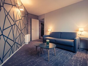 Une chambre d’hôtel moderne avec un canapé bleu, deux lampes de chevet, une petite table ronde et un mur orné d’un motif géométrique.