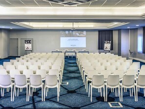 Un salón de conferencias con hileras de sillas blancas, una pantalla de proyección y un espacio para presentaciones.