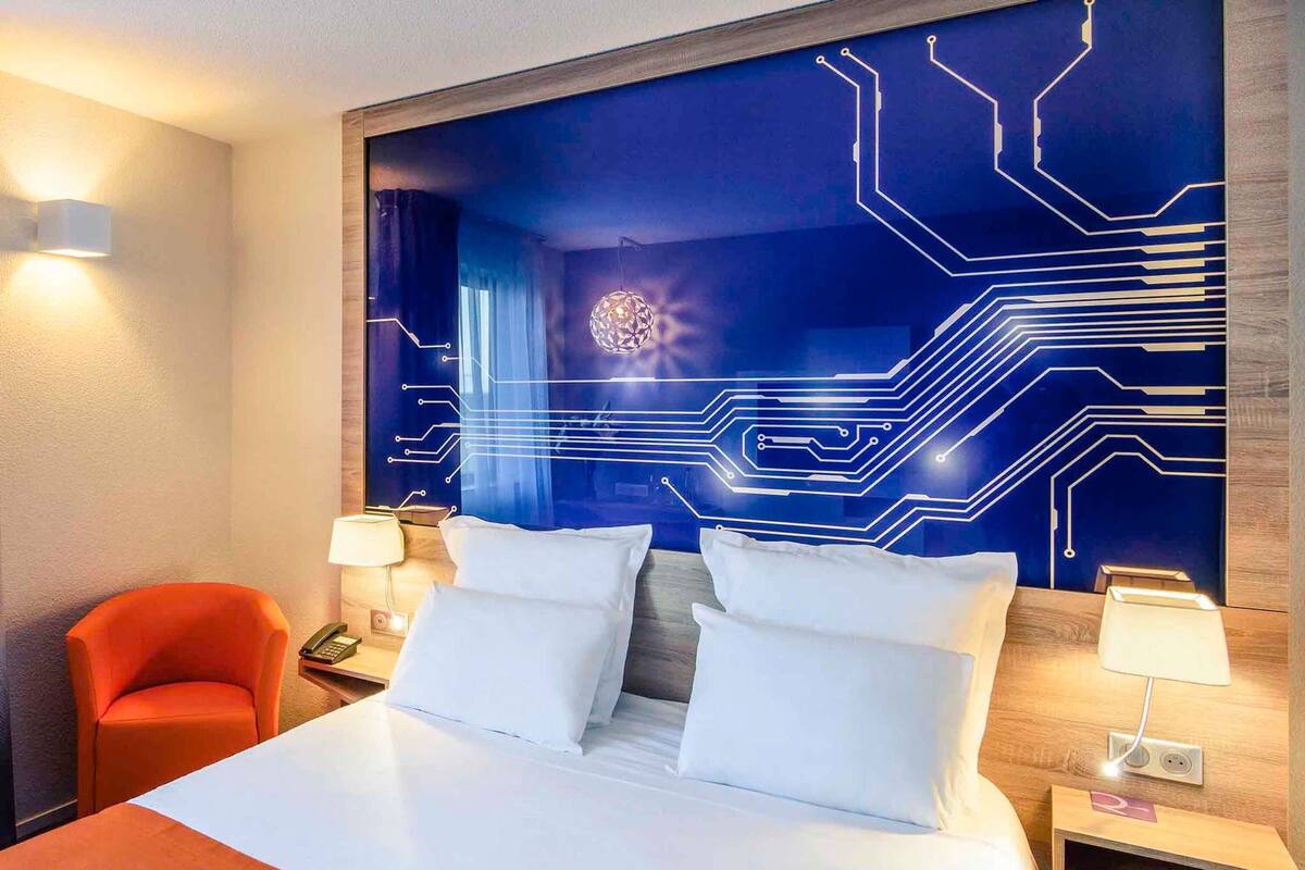 Ein Hotelzimmer mit einem Bett, zwei Nachttischlampen, einem Nachttisch, einem orangefarbenen Sessel und einer Wand mit einem blauen, an Schaltkreise erinnernden Muster.