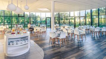 Un comedor amplio con mesas y sillas redondas, un mostrador para buffet y ventanas grandes que dan a zonas verdes.