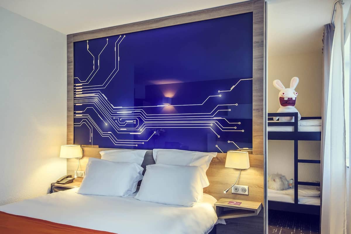 Une chambre d’hôtel avec un lit, des lampes de chevet, une table de chevet, un mur décoré avec un motif de circuit imprimé, une petite étagère avec un jouet en peluche et un lit superposé avec un jouet en peluche.