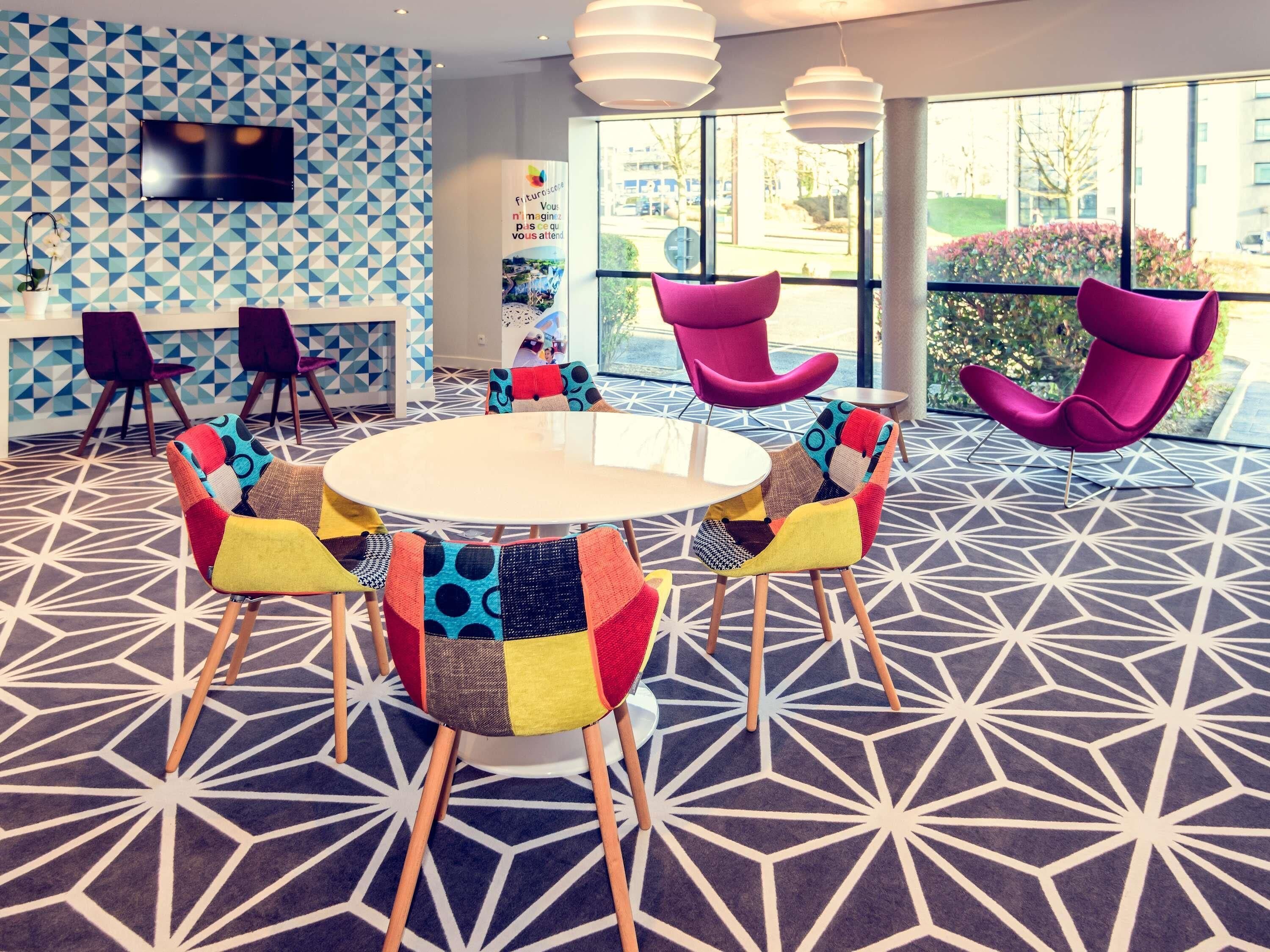 Une salle de conférence moderne, dotée d’une table ronde, de chaises aux couleurs vives, d’un tapis à motifs géométriques et de grandes fenêtres offrant une vue sur l’extérieur.