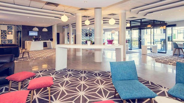 A modern lobby with a geometric patterned floor, seating areas with red and blue cushions, and a reception desk. - Mercure Poitiers Site du Futuroscope Hotel (Chasseneuil-du-Poitou)