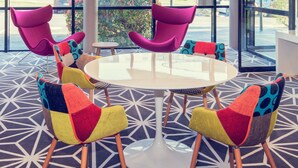 A modern indoor seating area with a round table and colorful chairs, large windows with a view of the street, and a geometric patterned floor. - Mercure Poitiers Site du Futuroscope Hotel (Chasseneuil-du-Poitou)