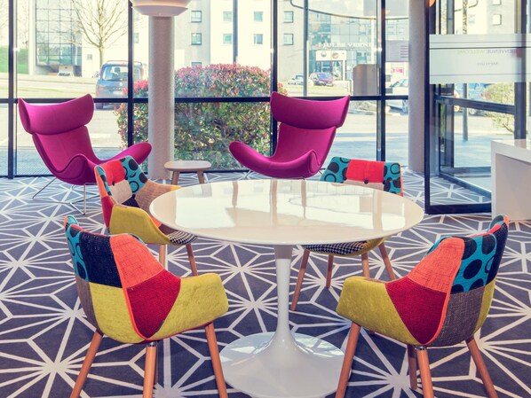 Un área de estar interior moderna con una mesa redonda y sillas de colores, ventanas amplias con vista a la calle y un suelo con estampado geométrico.