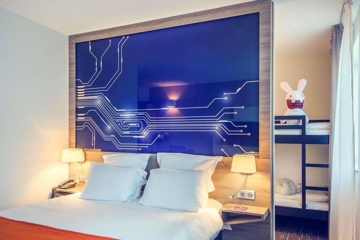 Ein Hotelzimmer mit Bett, Nachttischlampen, einem Nachttisch, einer Wand mit einem verzierungsartigen Muster und einem kleinen Regal mit einem Stofftier.