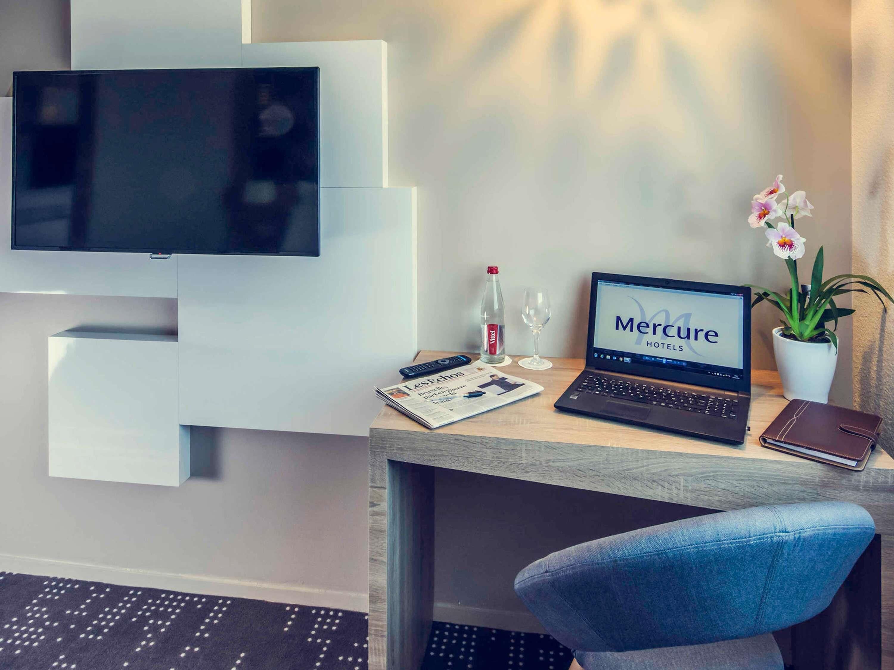 Habitación de hotel con escritorio, computadora portátil, control remoto, periódico, botella, copa de vino y planta de orquídea.