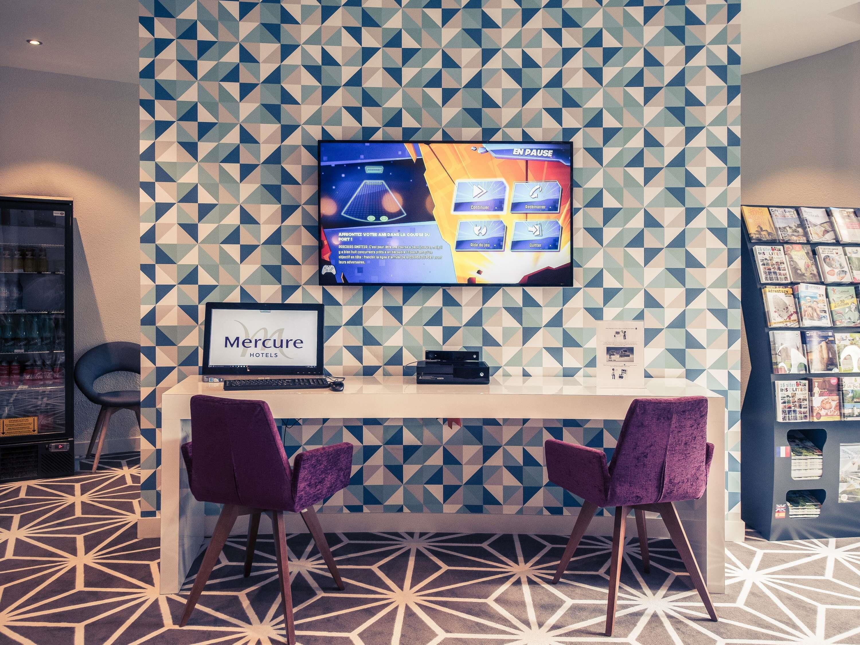 Un área de recepción con un moderno patrón geométrico en la pared, un televisor de pantalla plana mostrando un juego, un escritorio con una computadora portátil, dos sillas moradas y un perchero para revistas.