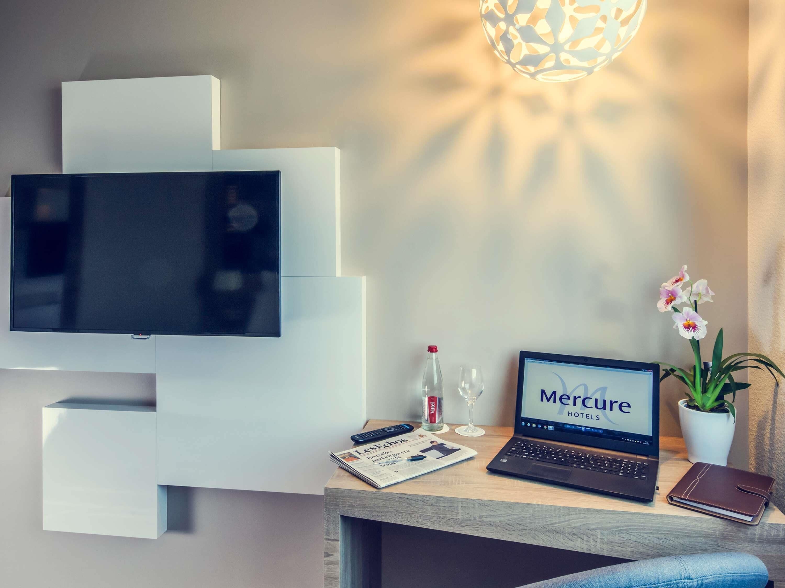 A modern hotel room with a flat-screen TV, a laptop, a magazine, a bottle, a glass, and a potted orchid.