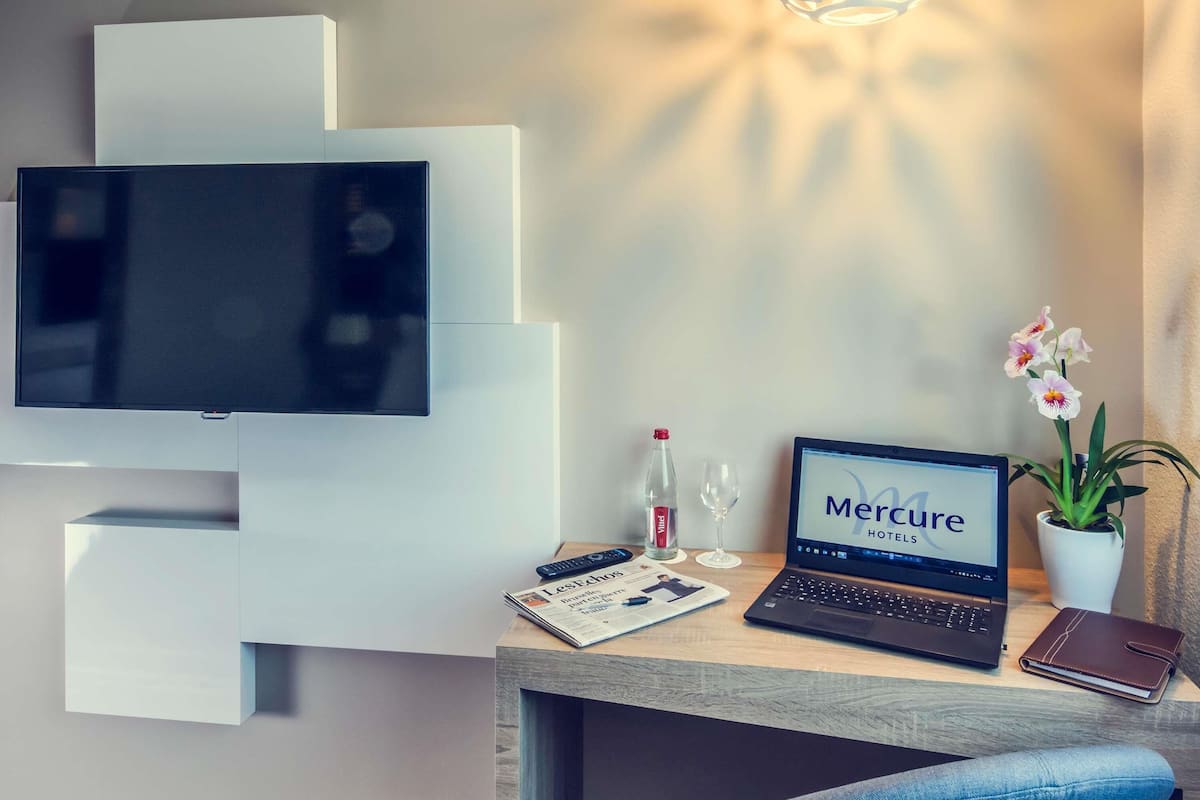 Une chambre d’hôtel moderne équipée d’un téléviseur à écran plat, d’un ordinateur portable, d’un magazine, d’une bouteille, d’un verre et d’une orchidée en pot.