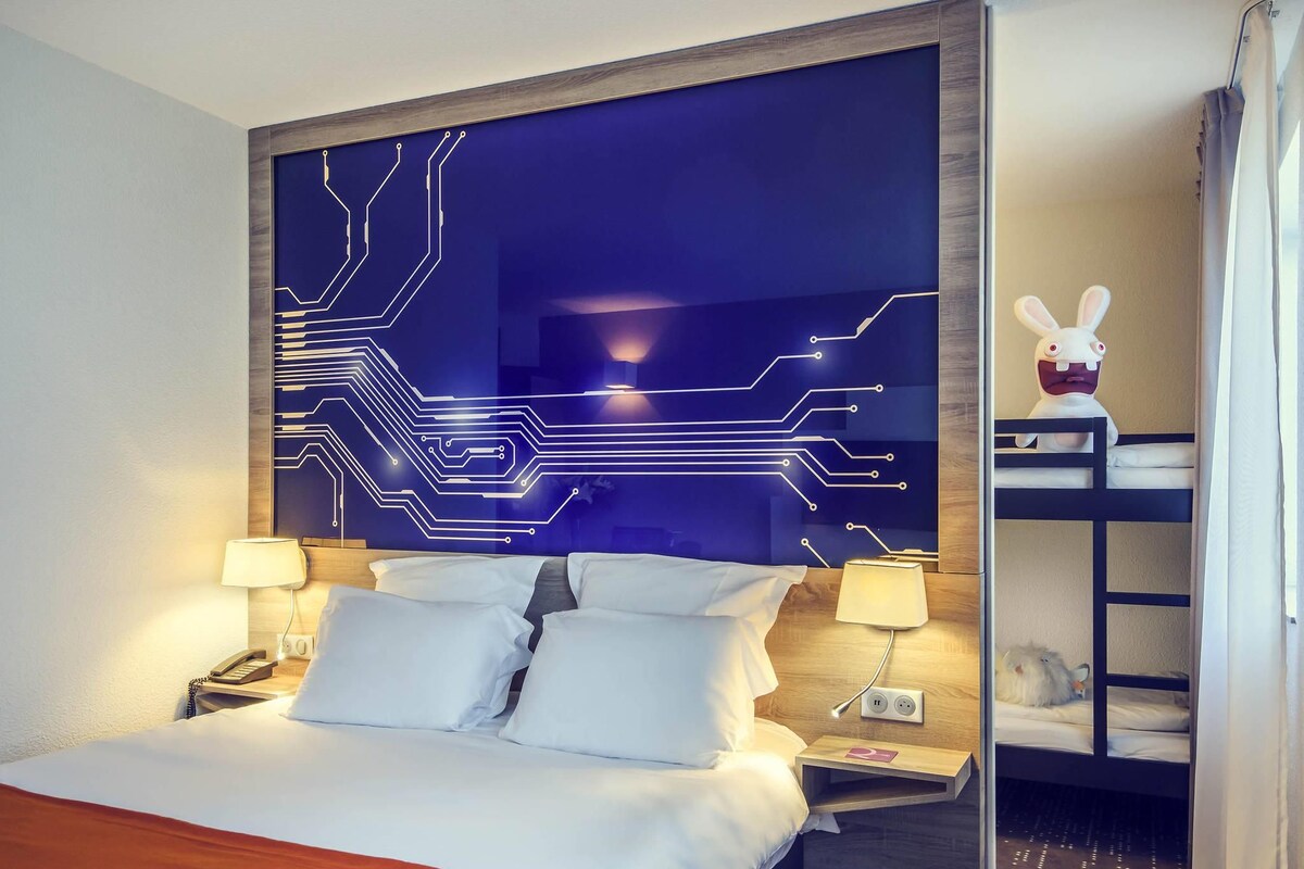 Une chambre d’hôtel avec un lit, des lampes de chevet, une table de chevet, un mur décoré avec un motif de circuit imprimé, une petite étagère avec un jouet en peluche et un lit superposé avec un jouet en peluche.