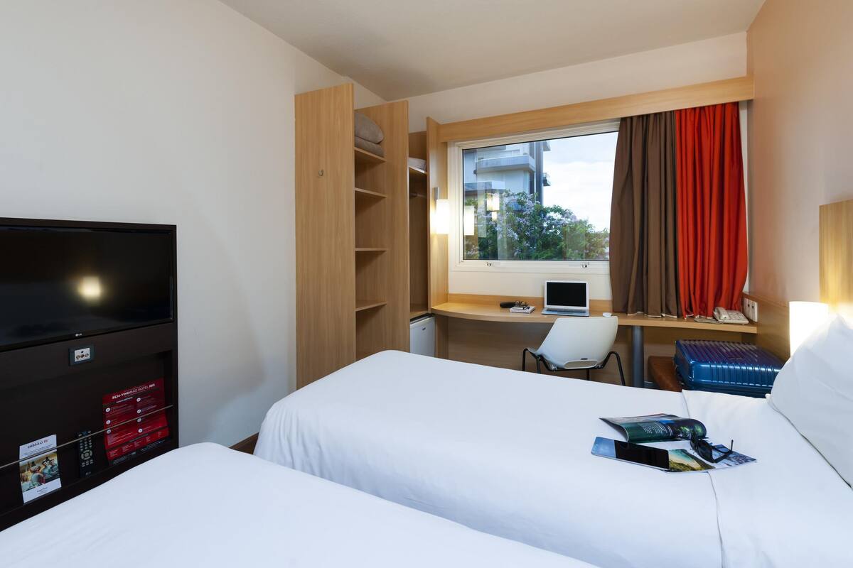 A hotel room with two beds, a TV, a desk with a laptop, a chair, and a window with curtains.