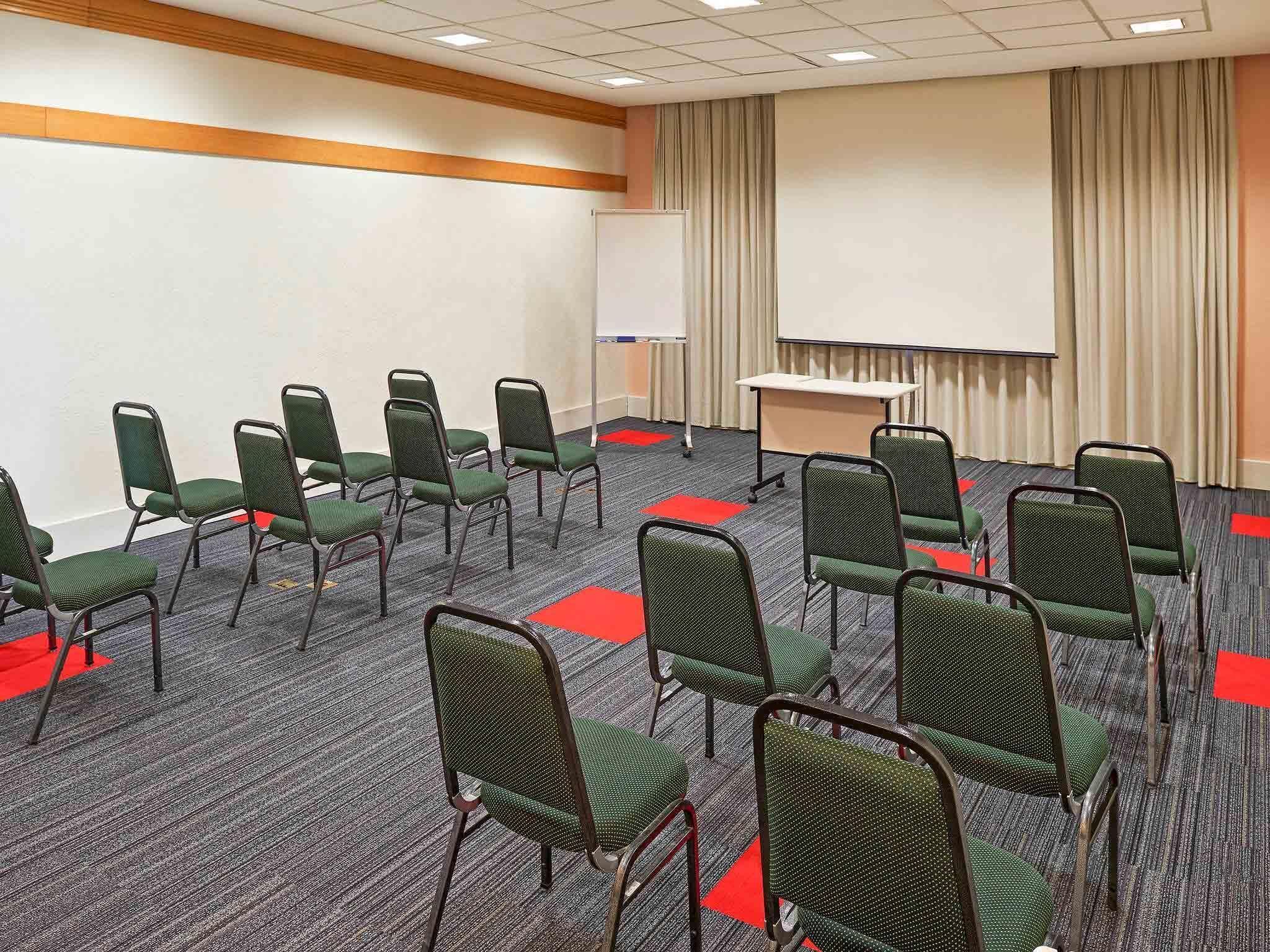 Uma sala de conferências com fileiras de cadeiras verdes, um quadro branco, uma tela de projetor e piso coberto de carpete.