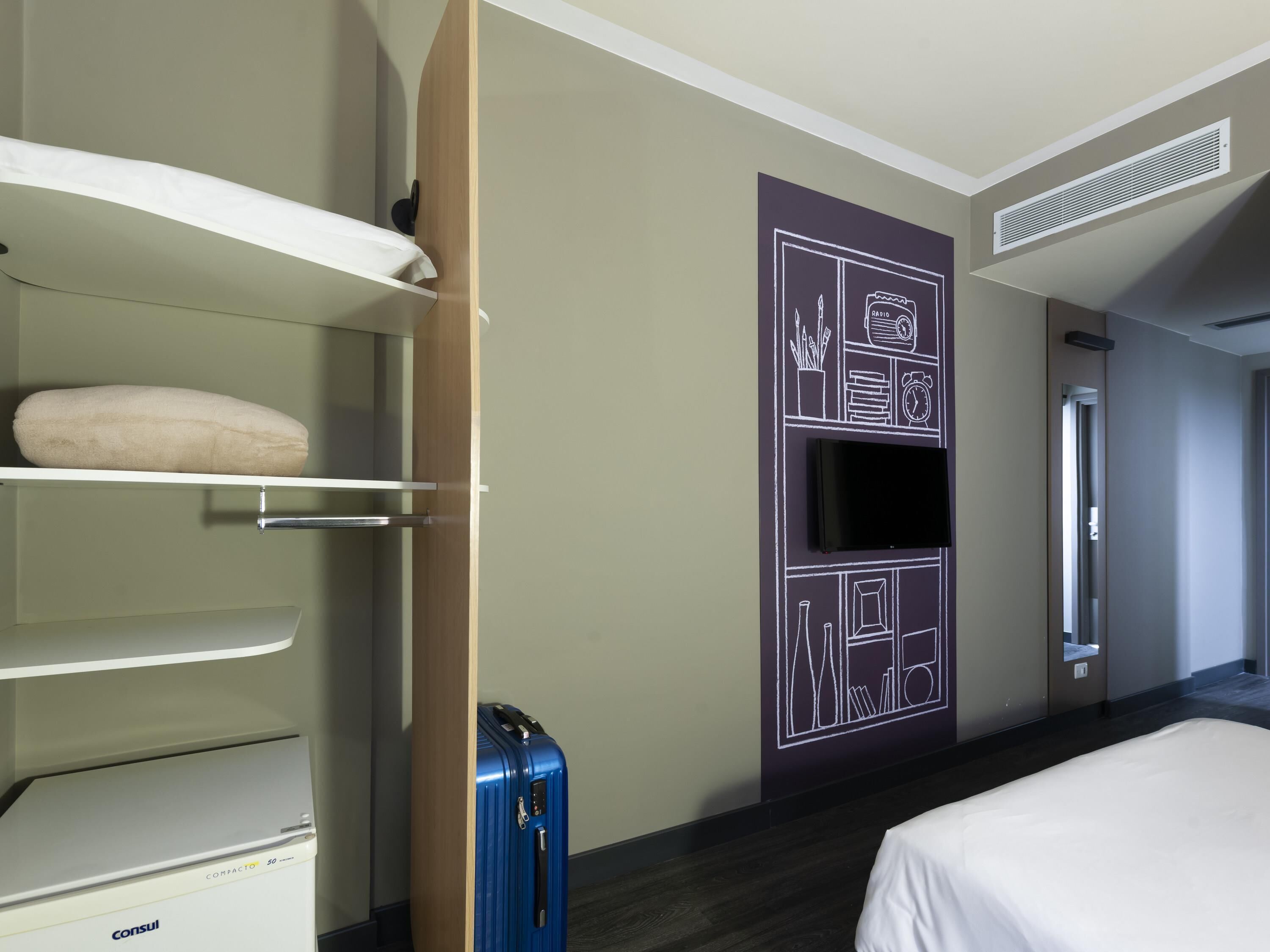 Een hotelkamer met een bed, een kledingkast met een opbergtas, een blauwe koffer en een aan de muur bevestigde televisie.