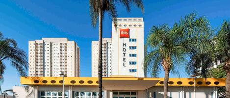 Un hotel de diseño moderno, con un llamativo letrero de 'ibis', una entrada central y palmeras alrededor.