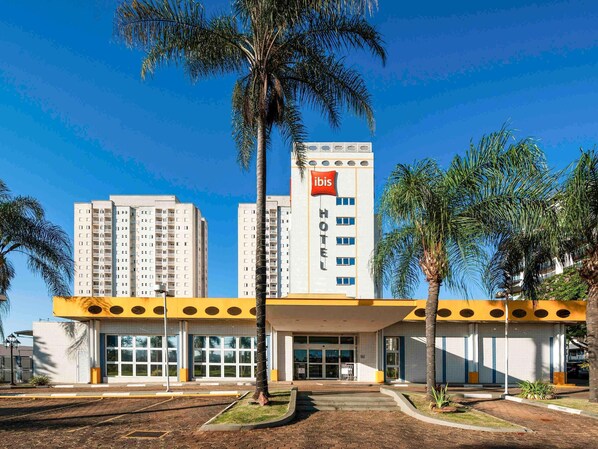 A hotel with a modern design, featuring a prominent 'ibis' sign, a central entrance, and surrounding palm trees. - ibis Sao Carlos (São Carlos)