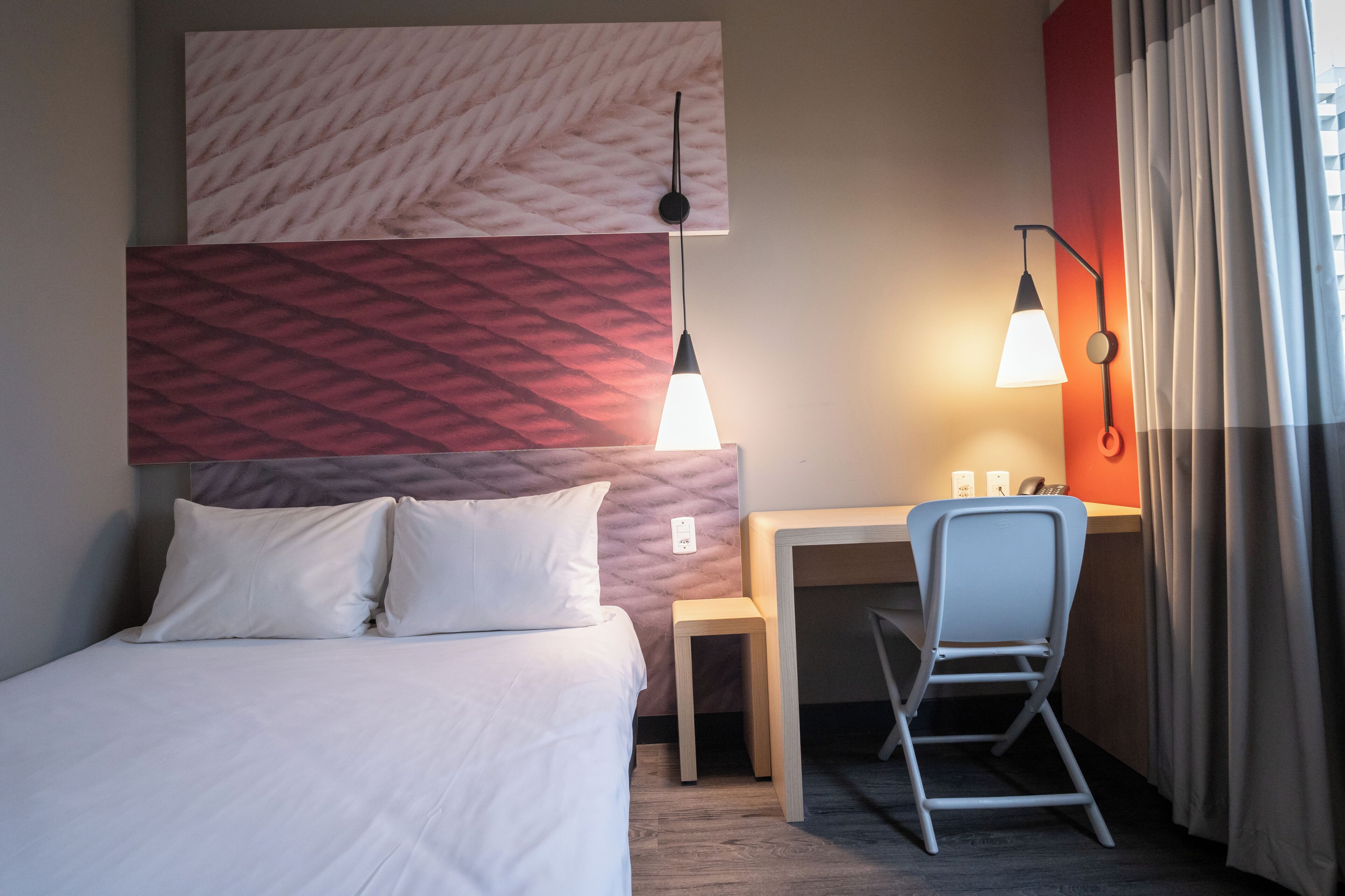 A hotel room with a bed, a desk, a chair, and a wall-mounted lamp.