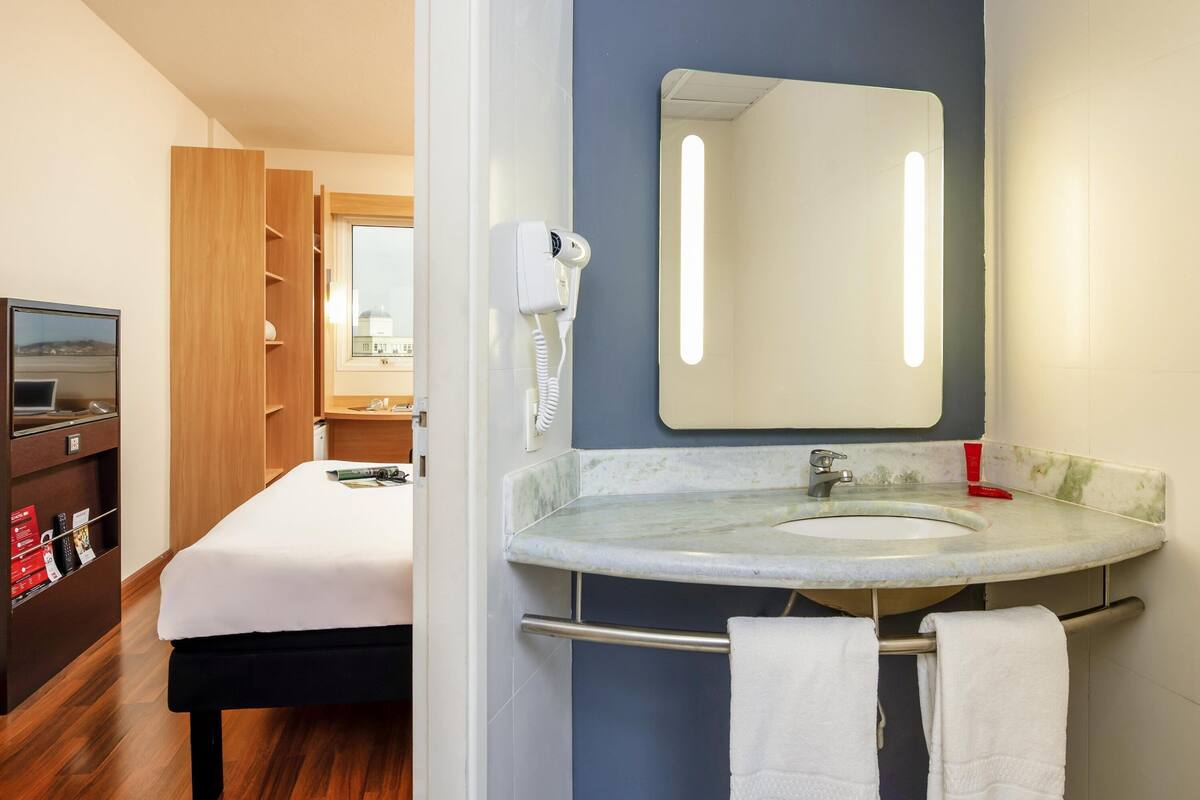 A hotel room with a bed, a desk with a laptop, a hairdryer, a sink with a mirror, and a window with a view of a building.